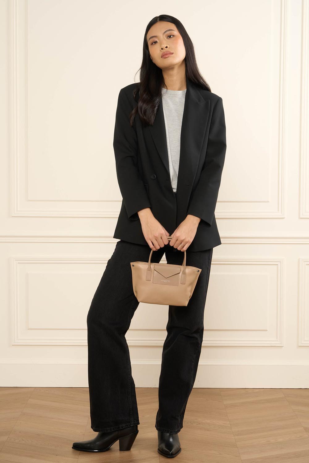 Lancaster, Maya KBA mini bag held in both hands, with a black blazer and trousers and a grey top.