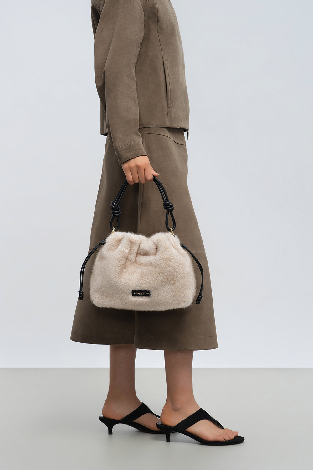 Lancaster Schoudertas met trekkoord M Moumoute Bruin – De beige Moumoute-schoudertas wordt in de hand gedragen en past bij een taupekleurige outfit en zwarte sandalen.