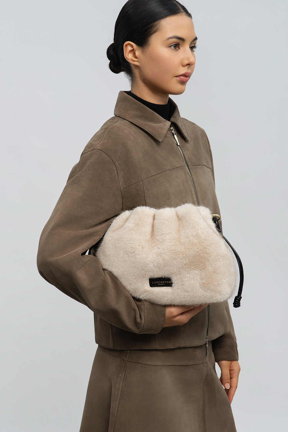 Lancaster Schoudertas met trekkoord M Moumoute Bruin – De Moumoute-schoudertas wordt met beide handen vastgehouden voor een bruin jasje en een bruine rok.