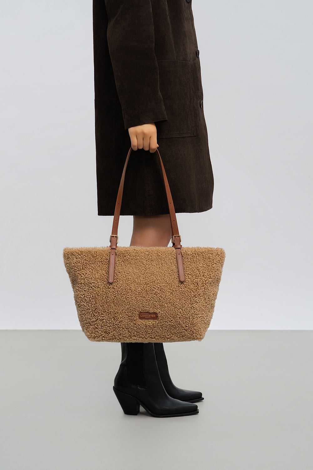 Lancaster, cabas Moumoute marron, porté à la main avec un manteau brun et des bottes noires.