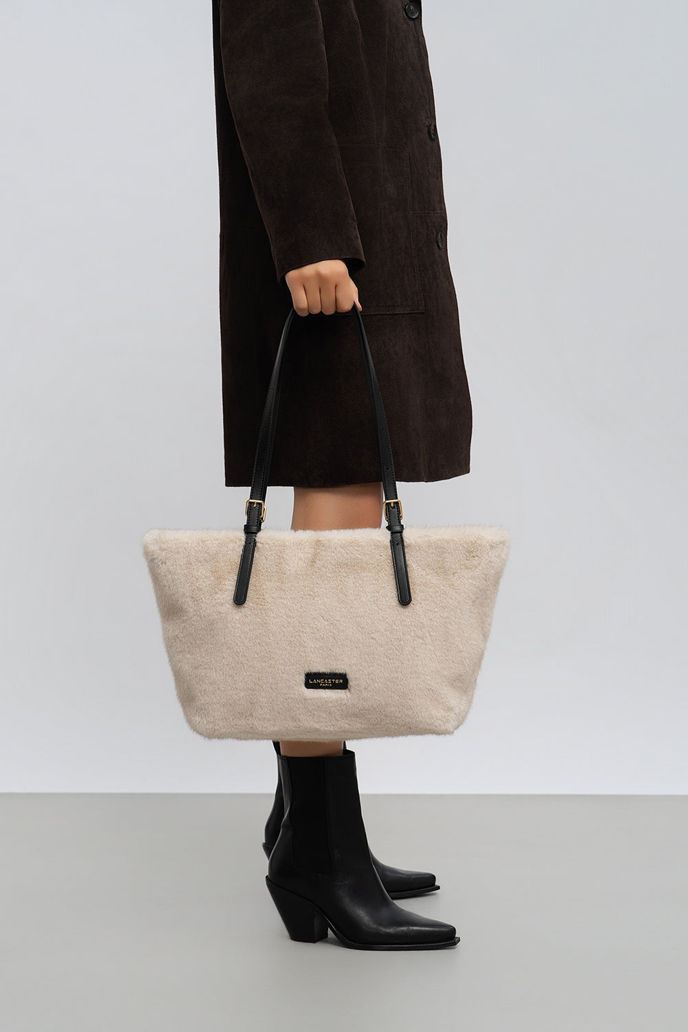 Lancaster cabas Moumoute beige porté à la main avec un manteau foncé et des bottes noires.
