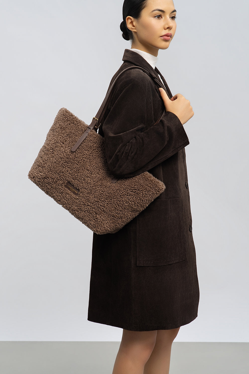 Lancaster, cabas Moumoute marron, porté à l’épaule avec un manteau brun foncé et une jupe noire.