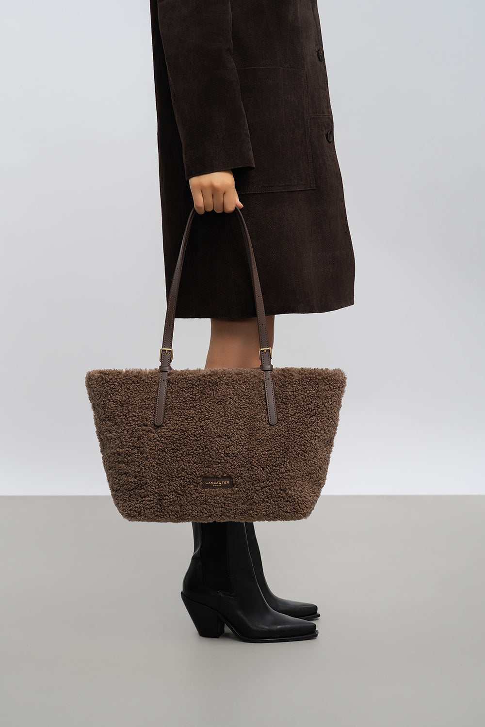 Lancaster cabas Moumoute marron porté à la main avec un manteau foncé et des bottes noires.