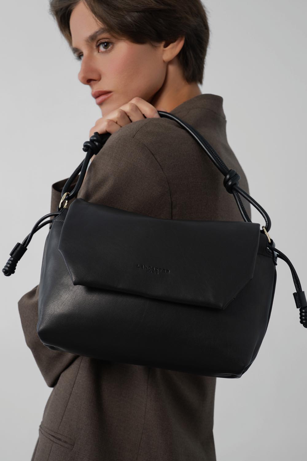 Lancaster Bandolera con solapa magnética M Lush Negro – El bolso bandolera Lush negro se lleva al hombro con una chaqueta marrón y un pantalón a juego.