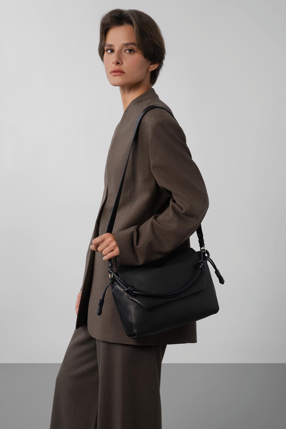 Lancaster Bandolera con solapa magnética M Lush Negro – En negro, el bolso bandolera Lush se lleva al hombro con un conjunto marrón de chaqueta y pantalón.