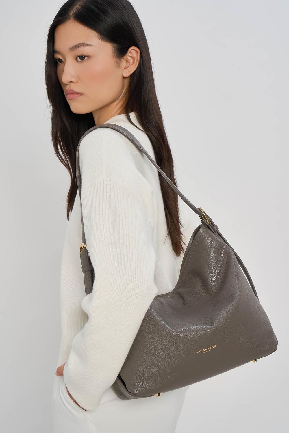 Lancaster Borsa a secchiello con zip Dune Blu – La borsa a secchiello Dune grigia si porta a spalla con un top bianco e pantaloni bianchi.