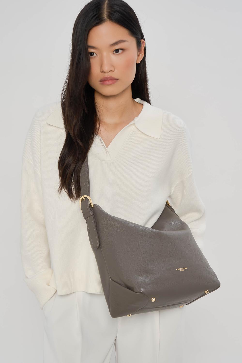 Lancaster Borsa a secchiello con zip Dune Blu – Con un top bianco e pantaloni bianchi, la borsa a secchiello Dune grigia è tenuta in mano.
