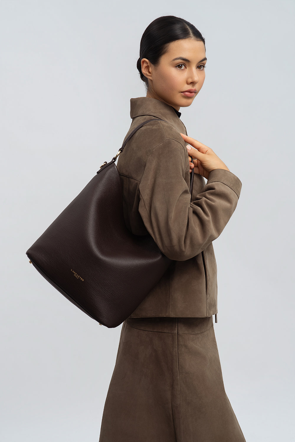 Lancaster Bolso cubo Con cremallera L Dune Borgoña Oscuro – El bolso saco Dune marrón va al hombro con una chaqueta y una falda marrones.