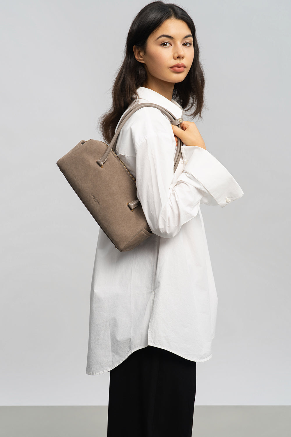 Lancaster Rectangle Bag Zippered Suède Cube Brown – The gray Rectangle bag is worn over the shoulder with a loose white shirt and black trousers.
