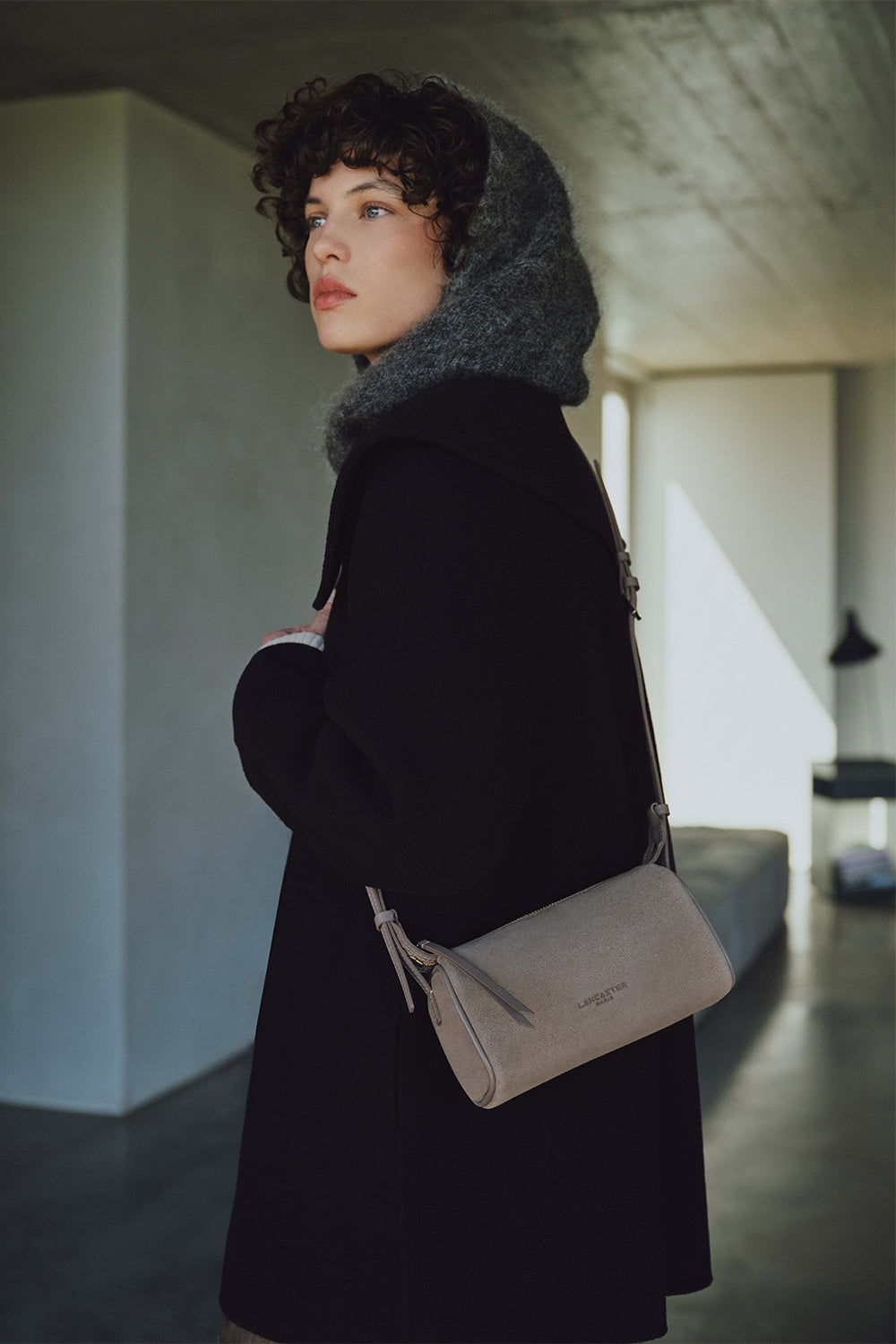 Lancaster, sac rectangle gris porté en bandoulière avec un manteau noir et une capuche grise.