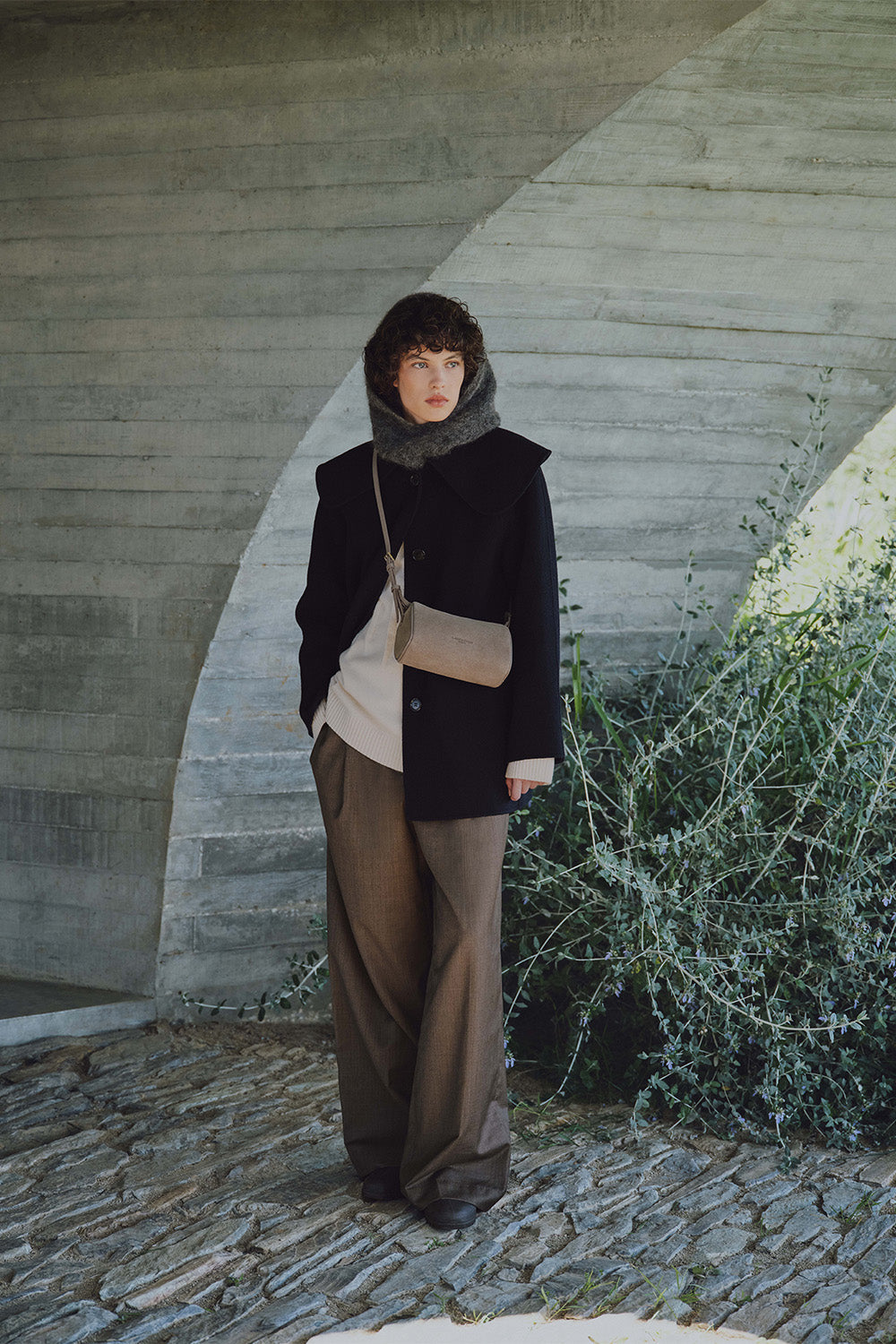 Lancaster, sac Rectangle gris porté en bandoulière avec un manteau noir, un haut beige et un pantalon marron.