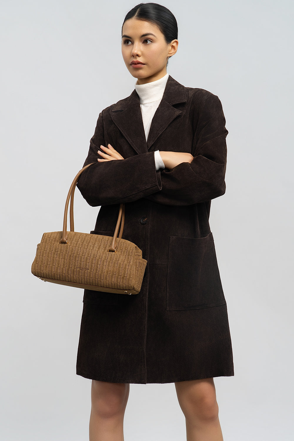 Lancaster rechteckige Tasche Tressé Cube in Braun, in der Hand getragen; dazu ein brauner Mantel und ein weißer Rollkragenpullover.