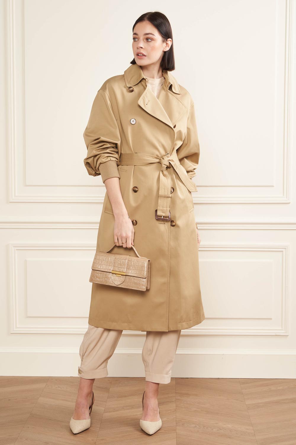 Lancaster Baguette-Tasche Exo Ily M Schwarz Krokoprägung – In der Hand ergänzt die beigefarbene Baguette-Tasche einen kamelfarbenen Trenchcoat und eine beige Hose.