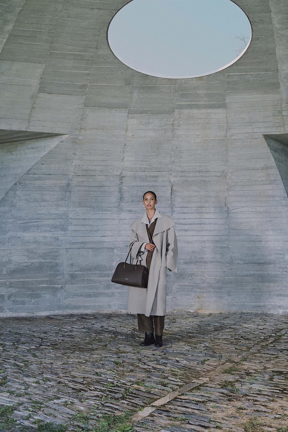 Lancaster, cabas Donna Hopper noir tenu à la main avec un long manteau gris et un pantalon sombre.