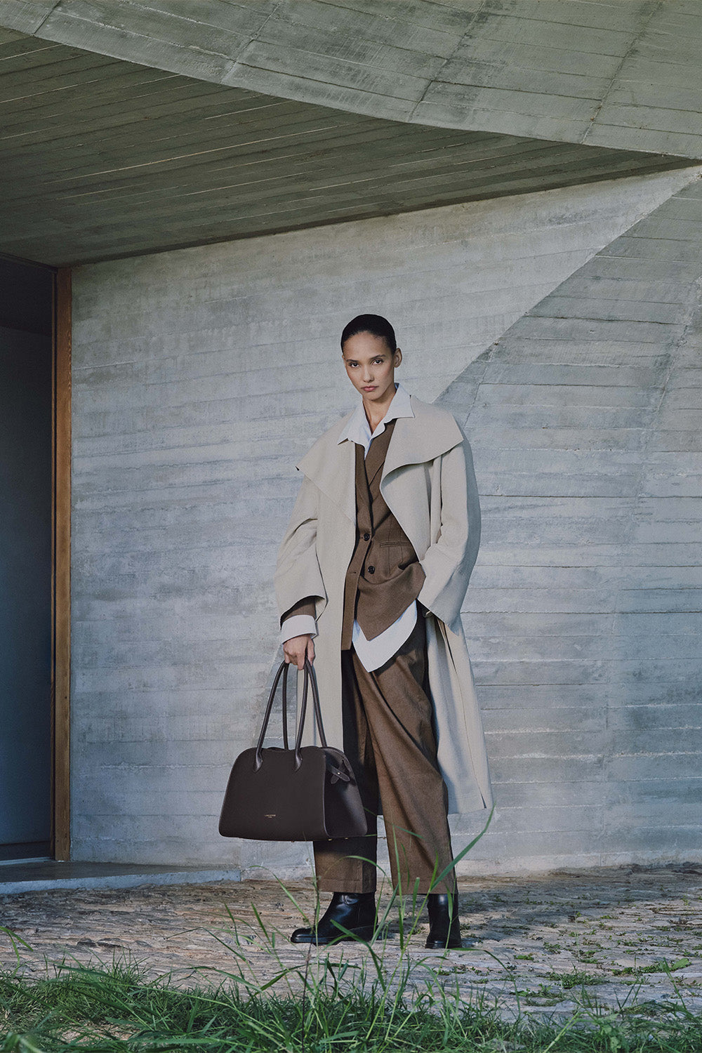 Lancaster, cabas Donna Hopper brun porté à la main avec un long manteau beige, une veste brune et un pantalon assorti.