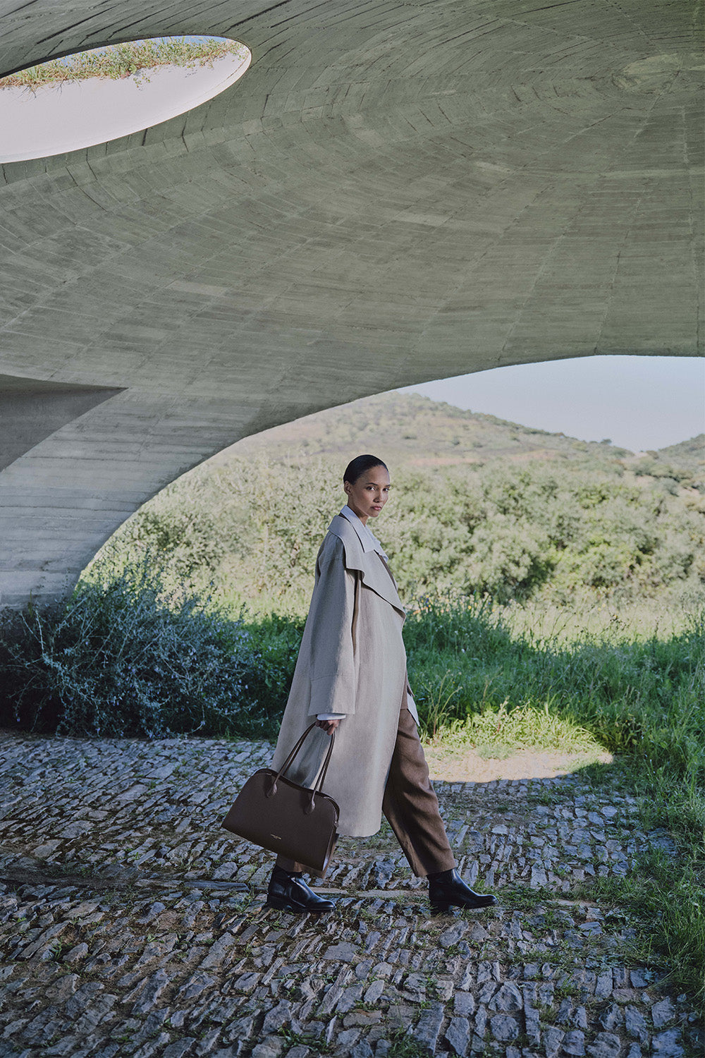 Lancaster, cabas Donna Hopper marron, tenu à la main avec un long manteau beige, un pantalon marron et des bottines noires.