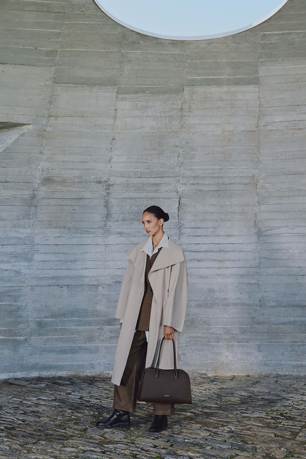 Lancaster, cabas Donna Hopper marron porté à la main avec un long manteau beige et une tenue sombre.