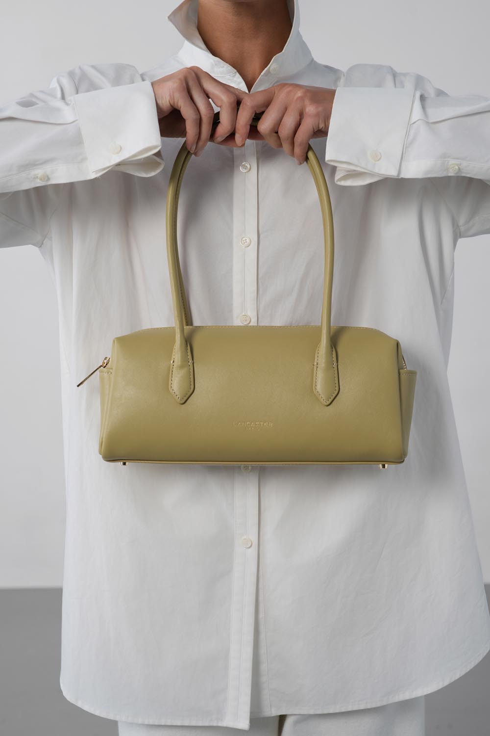 Lancaster Donna Cube Zipped Crossbody Bag Linen – The Donna Cube trotteur bag is held by hand in front of a loose white shirt.