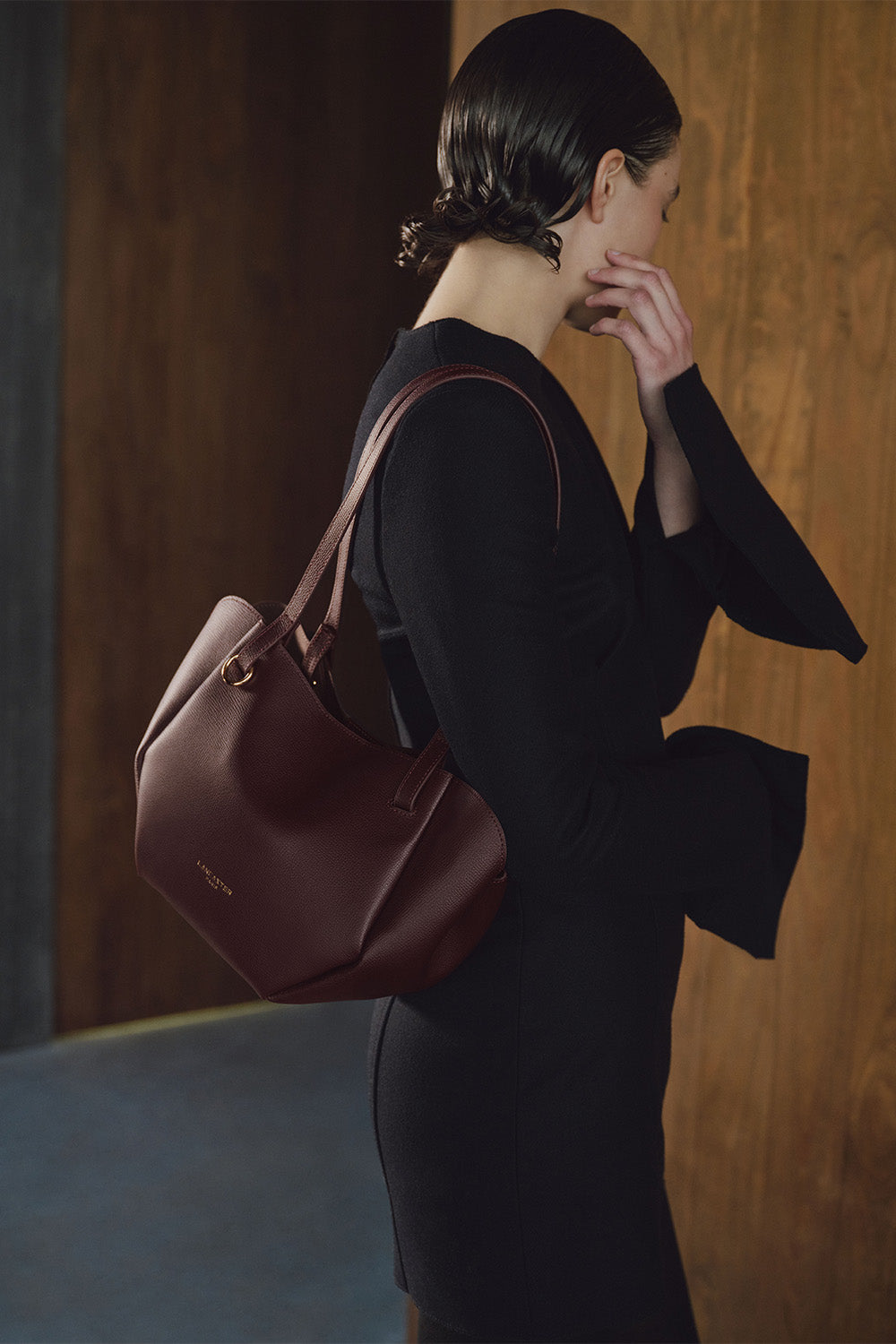 Lancaster Bolso Tote Sierra Haus Caqui – Llevado al hombro, un bolso burdeos acompaña un vestido negro de manga larga.