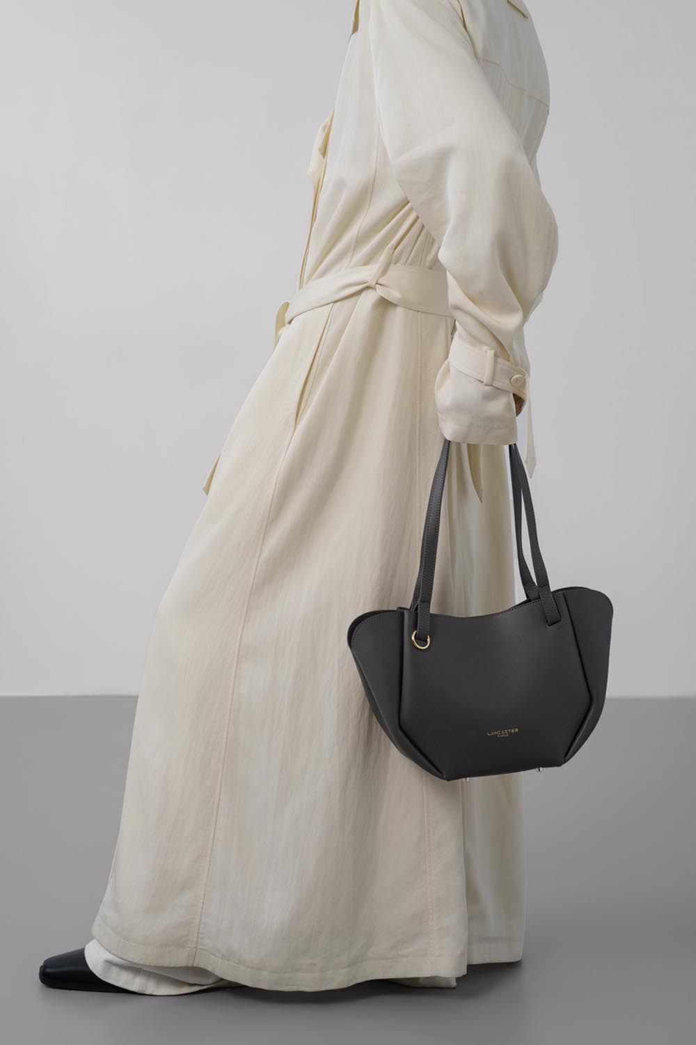 Lancaster Bolso Tote Sierra Haus Caqui – Llevado en la mano, el bolso acompaña un vestido largo blanco y zapatos negros.