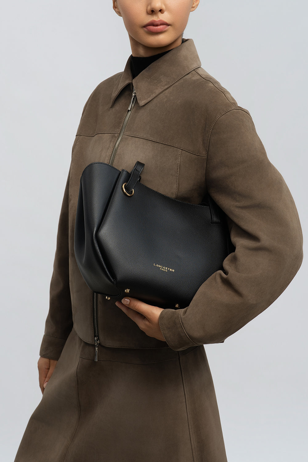 Lancaster Bolso Tote Sierra Haus Caqui – Llevado en el pliegue del brazo, el bolso negro acompaña a un vestido camisero marrón de manga larga.