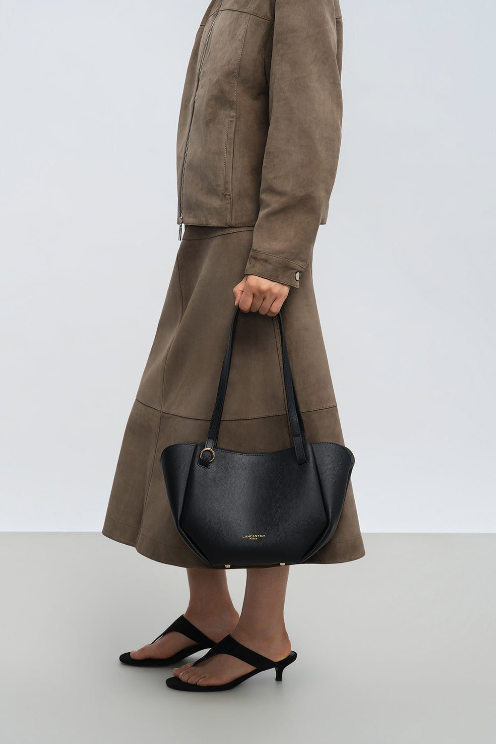 Lancaster Bolso Tote Sierra Haus Caqui – Llevado en la mano, con una chaqueta color topo, una falda acampanada a juego y sandalias negras.
