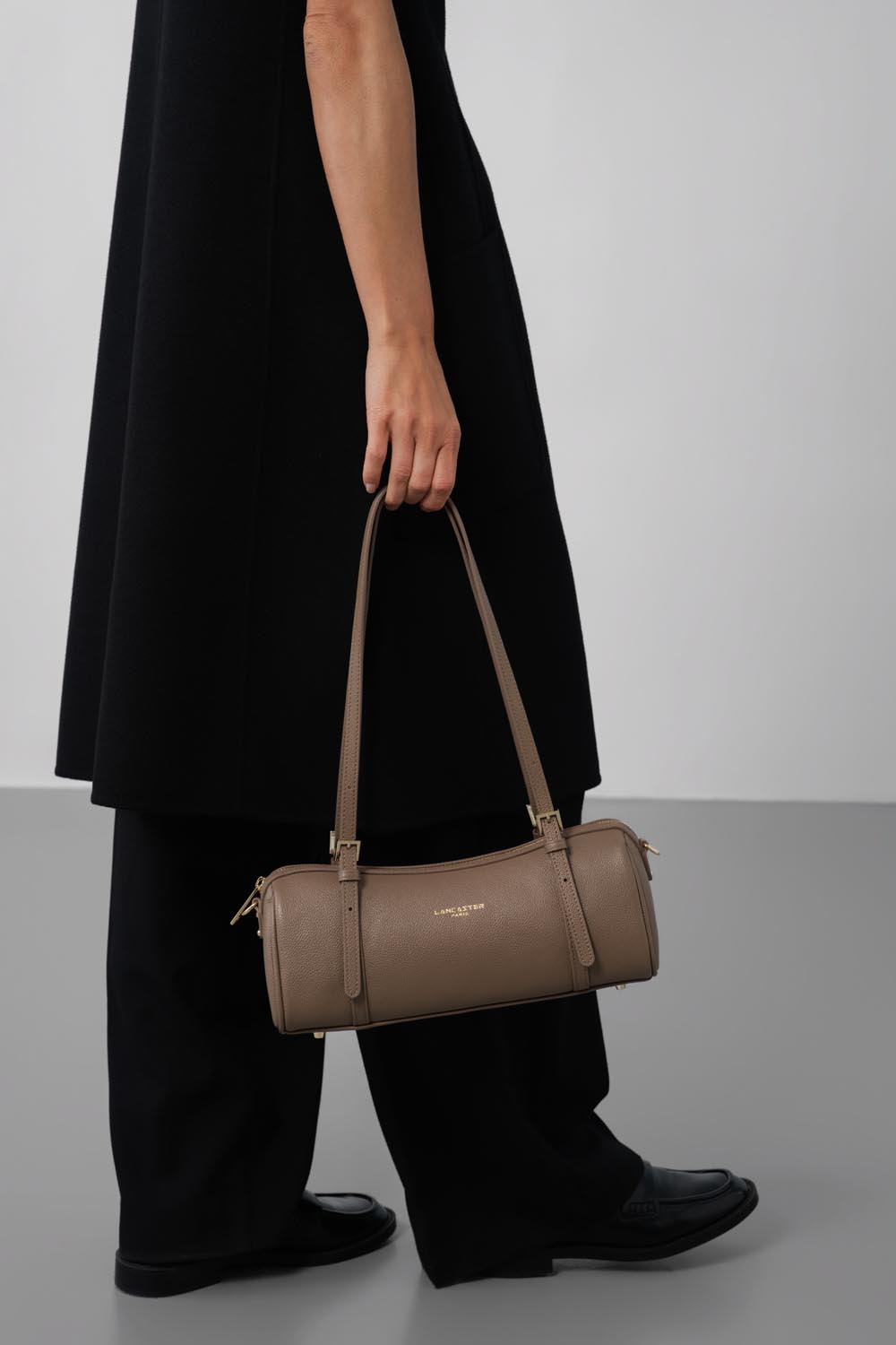 Lancaster Axelväska Med dragkedja Milano Ana Brun Mörk – Den beige baguetteväskan Milano Ana bärs i handen till en svart klänning och matchande skor.