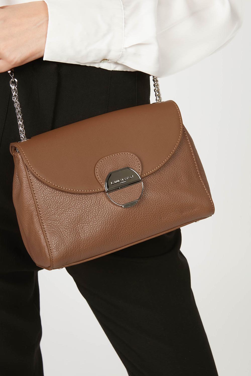 Lancaster Pia Camel Flap Crossbody Bag – The brown Pia Trotteur is worn crossbody with a white blouse and black trousers.