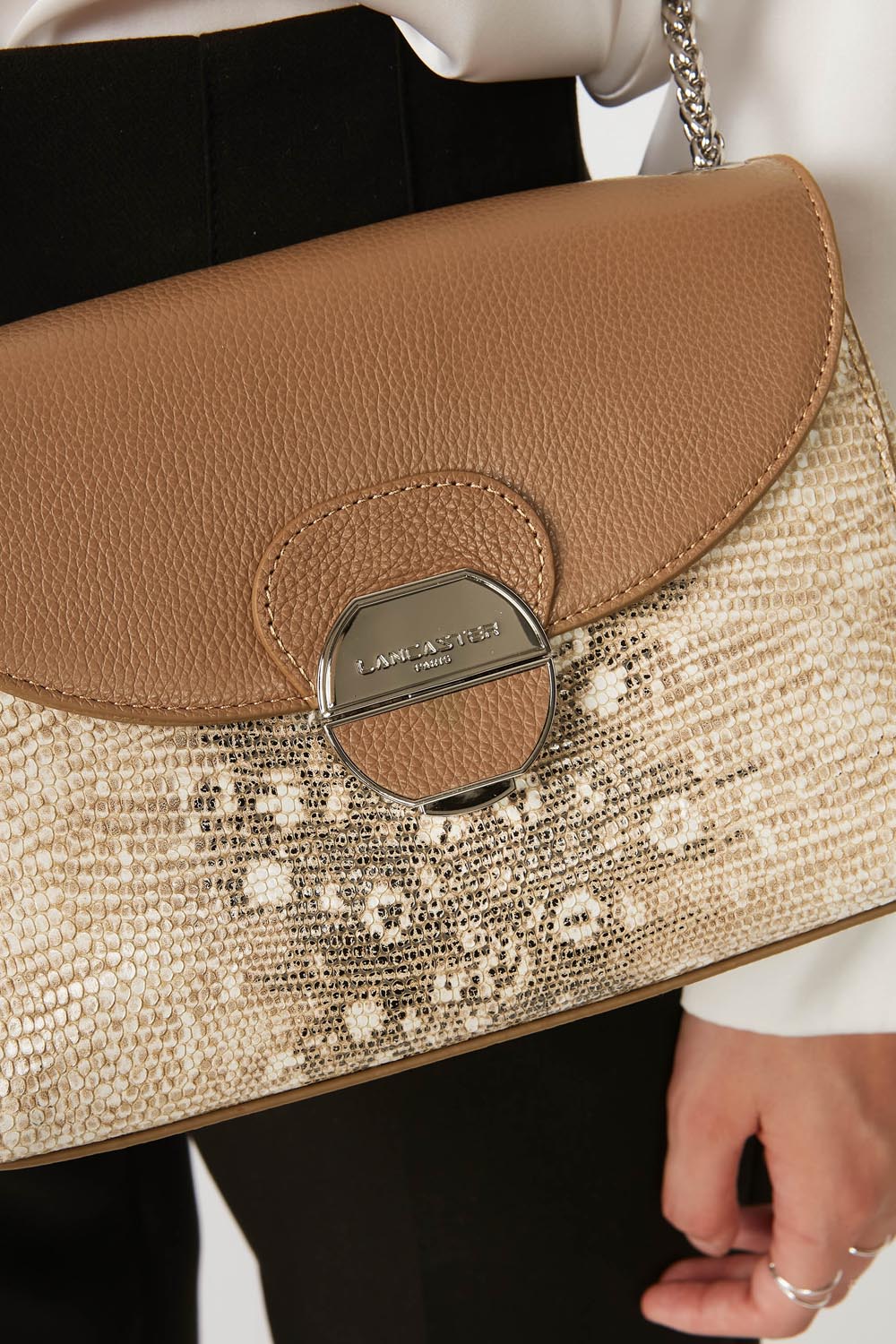 Lancaster Pia Camel Flap Crossbody Bag – The Pia satchel hangs from the shoulder with a black jacket and a white top.