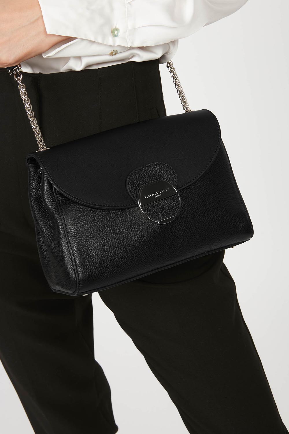Lancaster Pia Camel Flap Crossbody Bag – In black, the Pia crossbody bag is worn with a white shirt and black trousers.