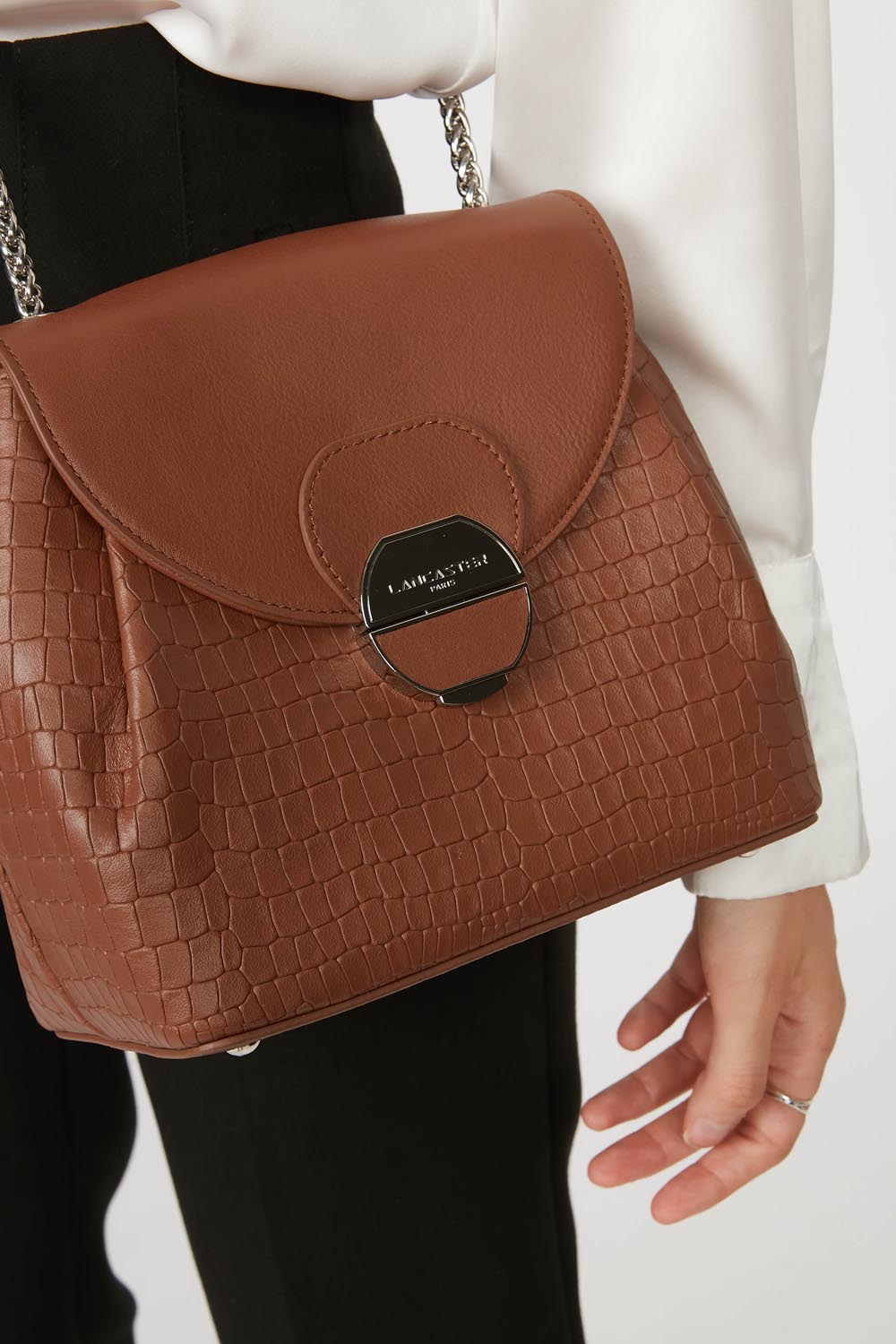 Lancaster Borsa a tracolla con patta e chiusura Pia Camel – La borsa Pia marrone è portata a spalla con un top bianco e pantaloni neri.