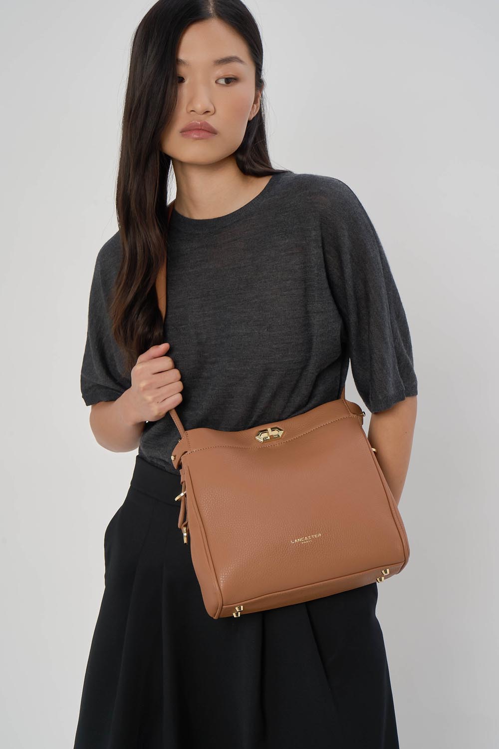 Lancaster Hobo-Tasche Mit Metallverschluss M Lady ROMY Camel – Zu einem grauen Oberteil und einem schwarzen Rock wird die Hobo Lady ROMY in der Hand gehalten.