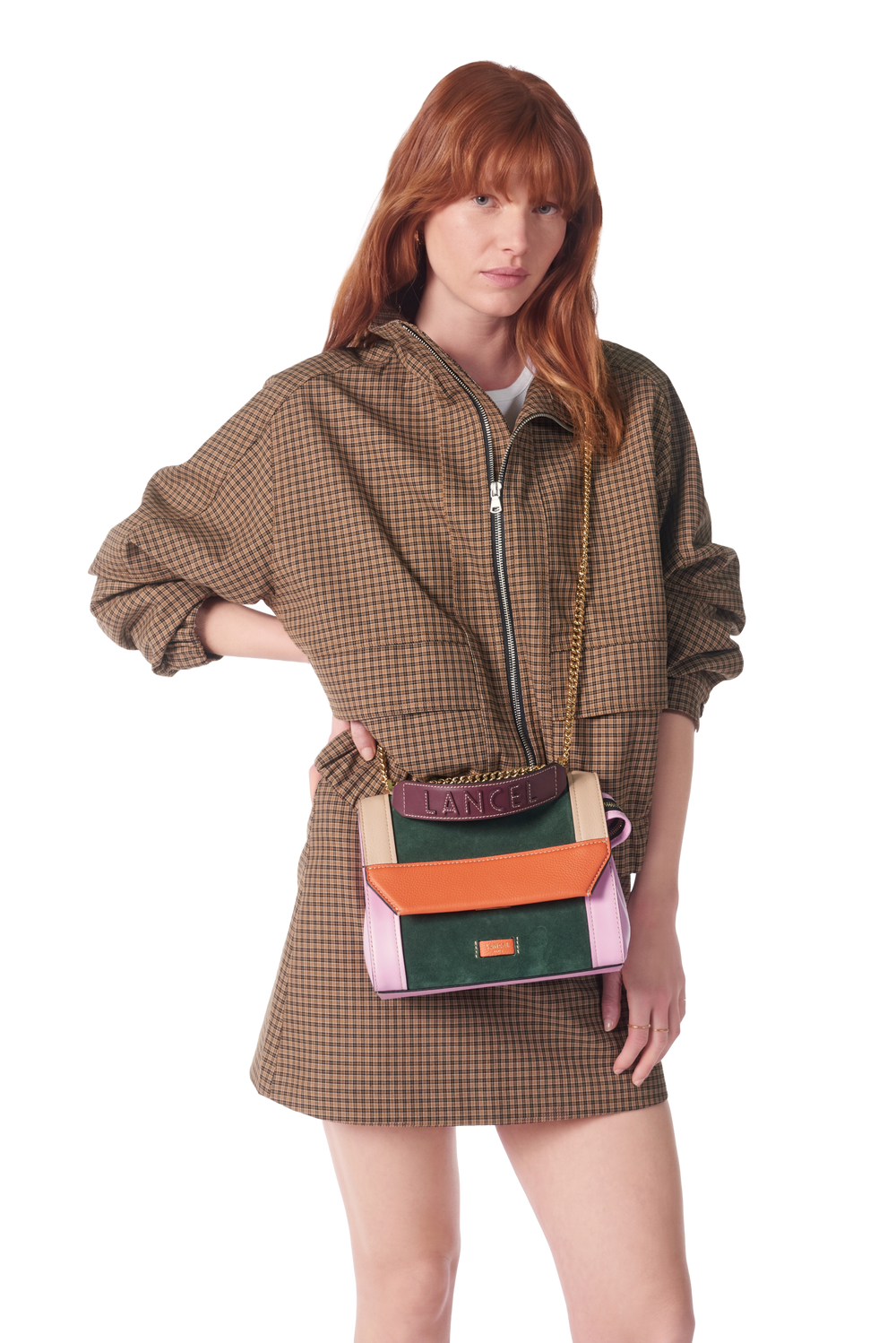 Lancel sac à rabat M Ninon vert porté en bandoulière avec une veste à carreaux bruns.