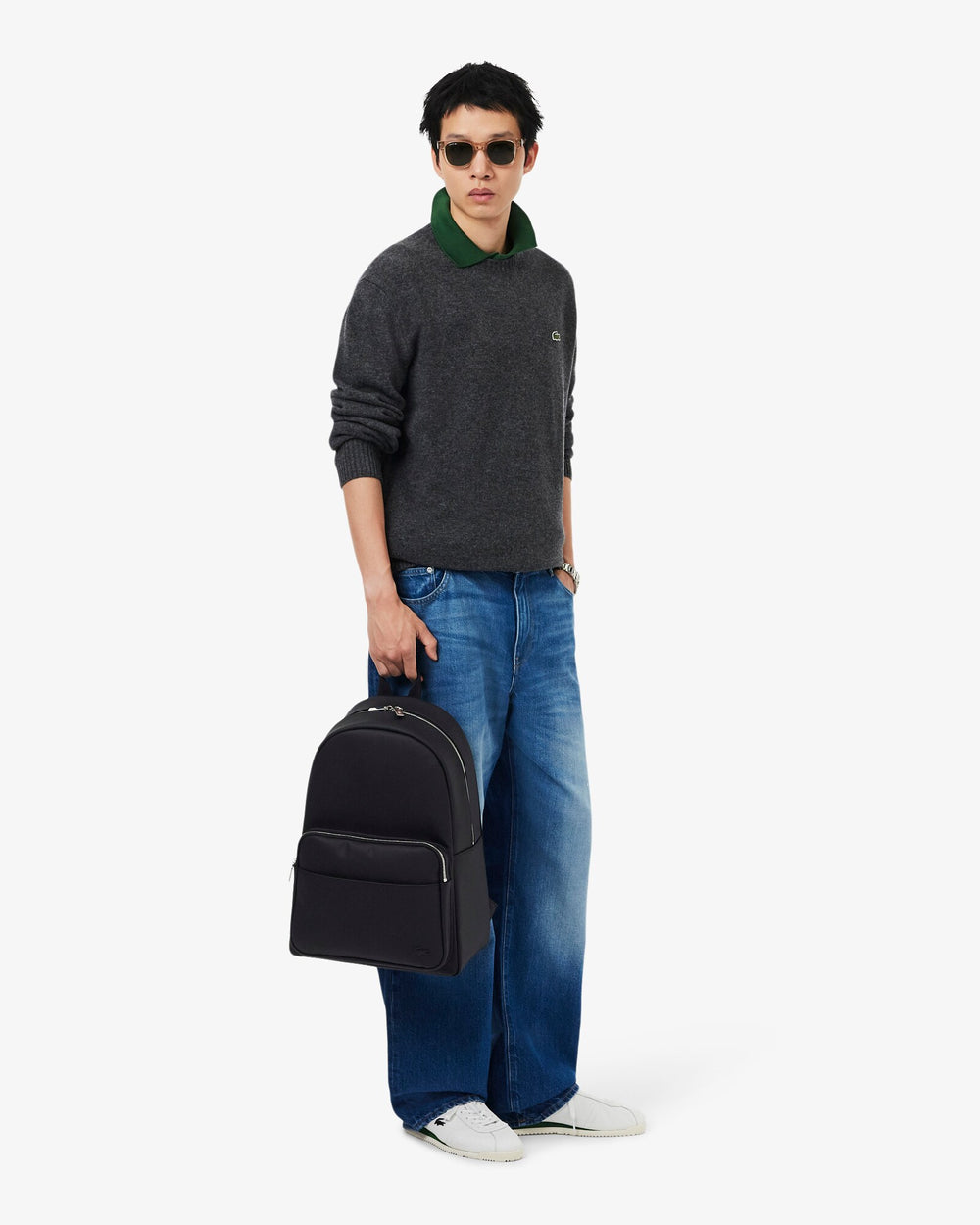 Lacoste Men’s Classic -reppu mustana kädessä, harmaan neuleen ja sinisten farkkujen kanssa.