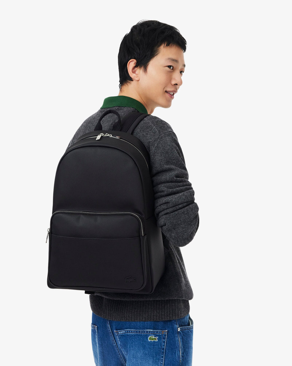 Lacoste musta Men’s Classic -reppu selässä, harmaan neuleen ja sinisten farkkujen kanssa.