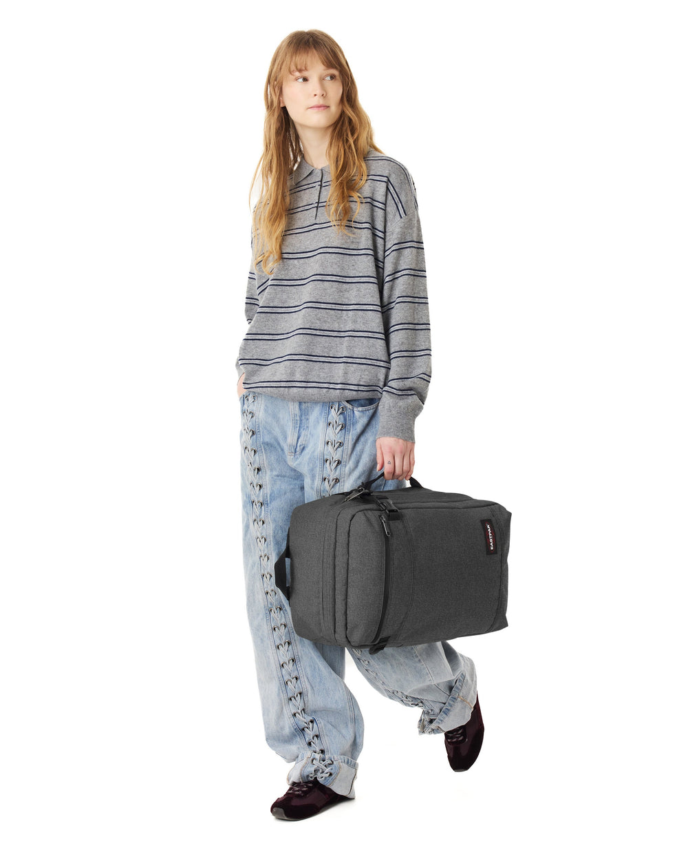 Eastpak szara torba podróżna Transit’r Pack trzymana w dłoni, z szarym pasiastym swetrem i jasnoniebieskimi dżinsami.