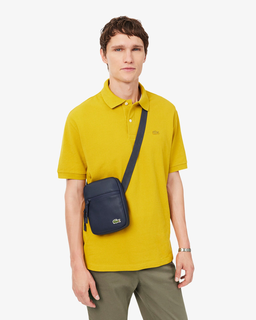 Lacoste borsa a tracolla Lcst blu, portata a tracolla con una polo gialla e pantaloni grigi.